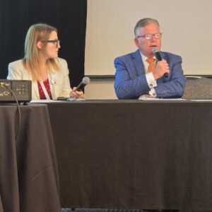 Panelists at a conference a man in a blue suit speaks into a handheld microphone as a woman in a white blazer sits beside him Unauthorized Practice of Law AAJ paralegal Brian Franciskato