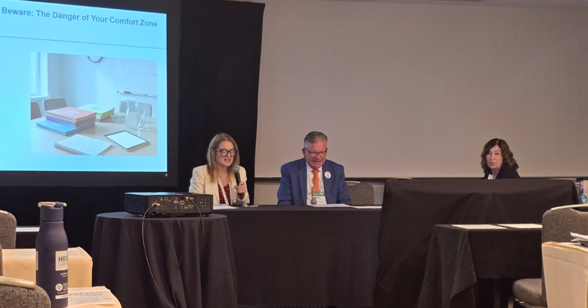 Three panel speakers at a conference table projector screen to the left showing a slide about the danger of your comfort zone