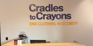 Reception area with large purple text Cradles to Crayons and orange END CLOTHING INSECURITY on a cream wall front desk visible in foreground indicating a charity or nonprofit organization Reception area with large purple text'Cradles to Crayons' and orange 'END CLOTHING INSECURITY' on a cream wall, front desk visible in foreground, indicating a charity or nonprofit organization.