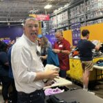 Man in a light checkered shirt smiles playfully at a clothing sorting event holding clothes near a table in a busy donation center
