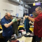 AAJ Service project   Two volunteers in a warehouse donation center sort clothes on tables with more people sorting in the background and yellow bins nearby