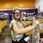Amanda Cryderman   Smiling staff member with glasses and a tattooed forearm folds a floral blouse at a clothing sorting area with yellow bins and a Sizing sign overhead